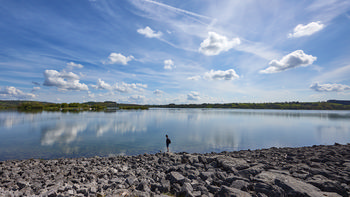 This landscape photograph captures Carsington Water, a large reservoir located on the edge of the Peak District, during a bright spring morning. The image features the calm lake reflecting scattered clouds across a blue sky, with the far shore showing green hills and patches of woodland typical of the region. The rocky shoreline in the foreground adds texture to the scene, and a lone figure stands near the water's edge, emphasizing the vastness of the reservoir. Known for its scenic views and recreational opportunities, Carsington Water is a prominent feature in the Peak District, often visited in spring for its tranquil setting and natural beauty.